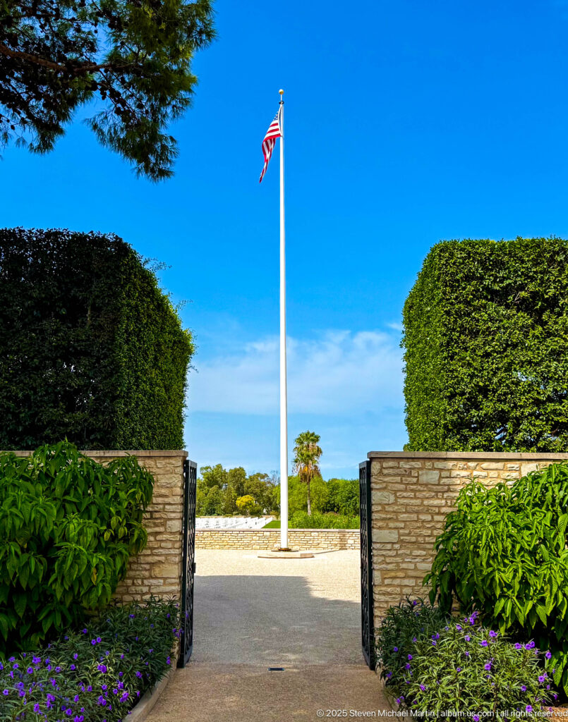 American flag on a tall white pole framed by clipped hedges, stone walls, flowers, and blue sky.