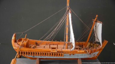 Wooden model of an ancient Mediterranean sailing ship with mast, rigging, curved bow, and long hull.