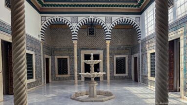 Enclosed courtyard with central tiered fountain, twisted columns, striped arches, tiled walls, grilled windows, and a marble floor.