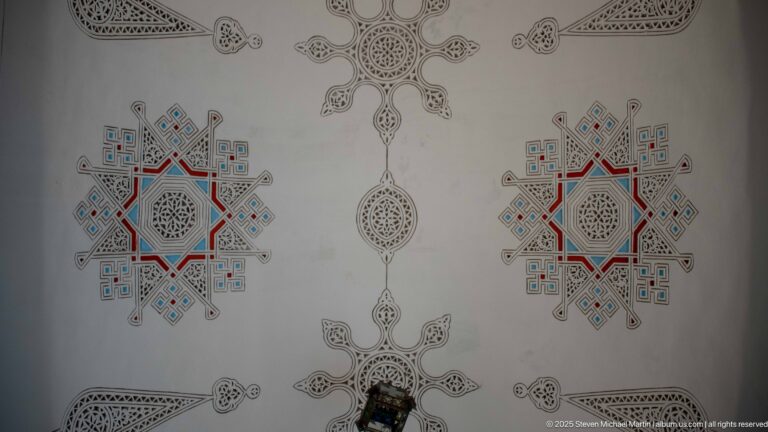 Pale wall with two matching red-and-blue star medallions, fine black geometric drawing, and pendant motifs along the center.