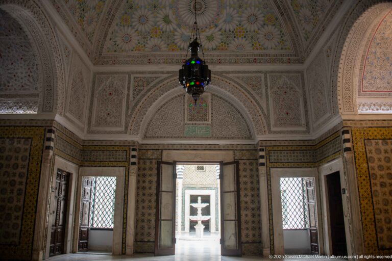 Vaulted palace room with floral ceiling ornament, hanging glass lantern, tiled walls, grilled windows, and an open doorway to the courtyard.