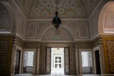 Vaulted palace room with floral ceiling ornament, hanging glass lantern, tiled walls, grilled windows, and an open doorway to the courtyard.