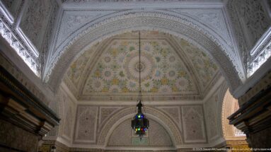 Polychrome semi-dome with radial floral ornament framed by white carved plaster and a dark multicolored lantern hanging below.