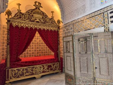 Ornate red canopy bed beside an inlaid wooden cabinet in a palace room with patterned tile walls and carved plaster above.