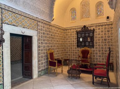 Tiled palace sitting room with red upholstered chairs, low tables, a dark latticed window, and three small colored-glass openings above.