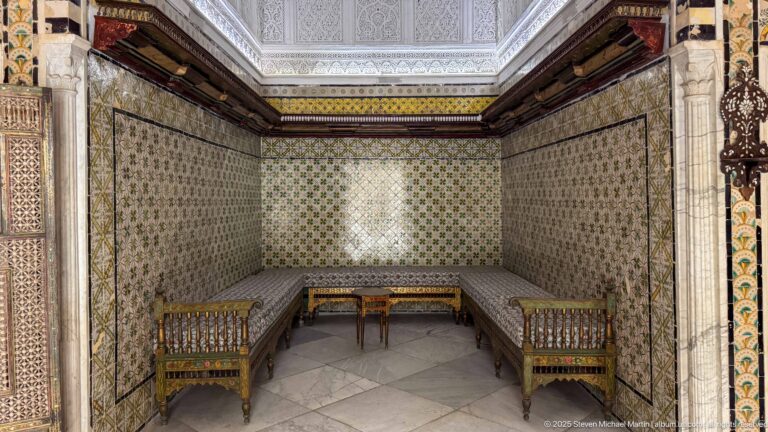 Small tiled alcove furnished with benches, two chairs, and a central table beneath richly patterned wall decoration.