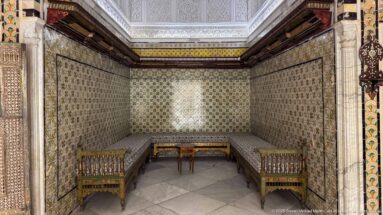 Small tiled alcove furnished with benches, two chairs, and a central table beneath richly patterned wall decoration.