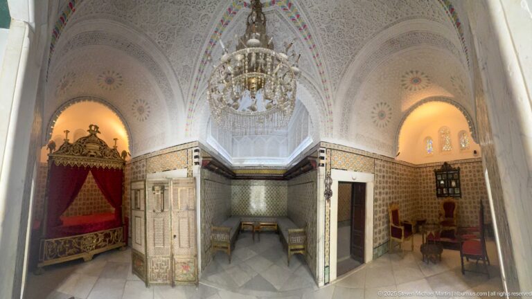 Ornate palace suite with red canopy bed, tiled walls, vaulted ceiling, chandelier, doors, chairs, and adjoining sitting alcoves.