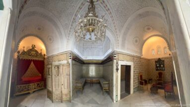 Ornate palace suite with red canopy bed, tiled walls, vaulted ceiling, chandelier, doors, chairs, and adjoining sitting alcoves.