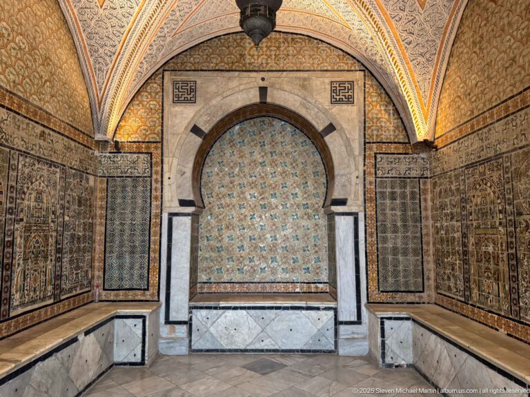 Symmetrical tiled chamber with a marble-framed horseshoe alcove, turquoise patterned back wall, and darker tile panels at both sides.