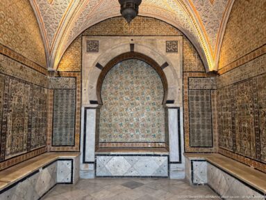 Symmetrical tiled chamber with a marble-framed horseshoe alcove, turquoise patterned back wall, and darker tile panels at both sides.