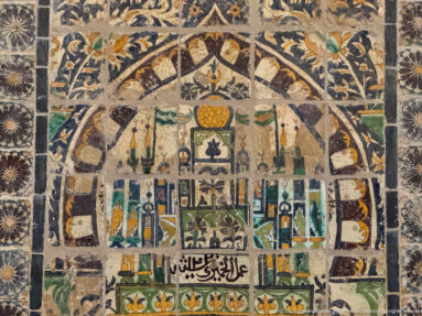 Polychrome tile panel with a central architectural motif, birds, flowers, vessels, and a horizontal Arabic inscription near the bottom.