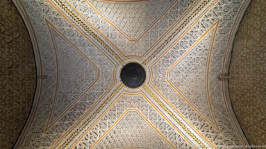 Four-part white carved-plaster vault outlined by gilded ribs and converging on a dark circular boss at the center.
