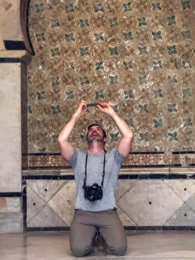 Kneeling male visitor holding a phone overhead beneath dense floral and geometric palace decoration, with a camera hanging from his neck.