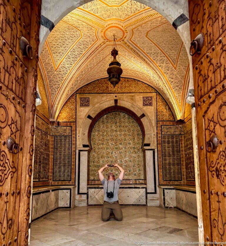 Kneeling visitor centered before a tiled horseshoe-arched alcove, framed by open decorated wooden doors and a vaulted ceiling.