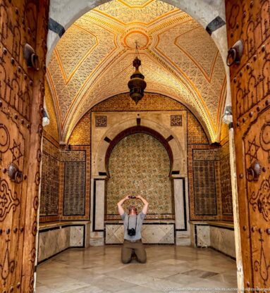 Kneeling visitor centered before a tiled horseshoe-arched alcove, framed by open decorated wooden doors and a vaulted ceiling.