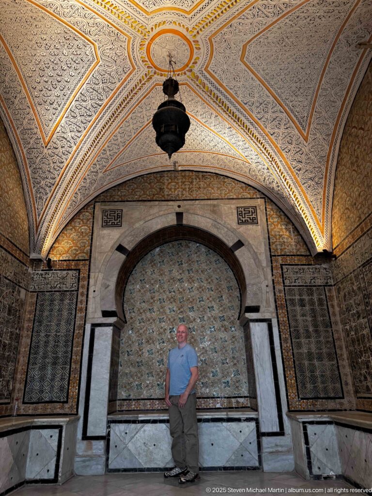 Standing visitor before a marble-framed horseshoe alcove with patterned tilework, flanking panels, and a centered hanging lantern.