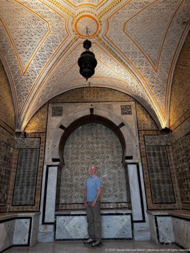 Standing visitor before a marble-framed horseshoe alcove with patterned tilework, flanking panels, and a centered hanging lantern.