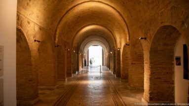 Long brick barrel-vaulted corridor with repeated side arches, warm spotlights, and a bright doorway at the far end.