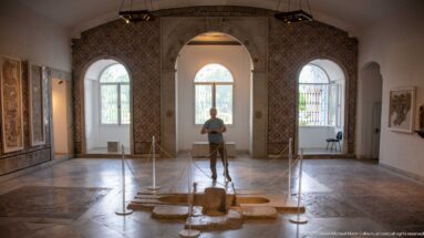 Roped low stone remains in the center of a gallery with a standing visitor, three tall arched windows, and patterned wall decoration.