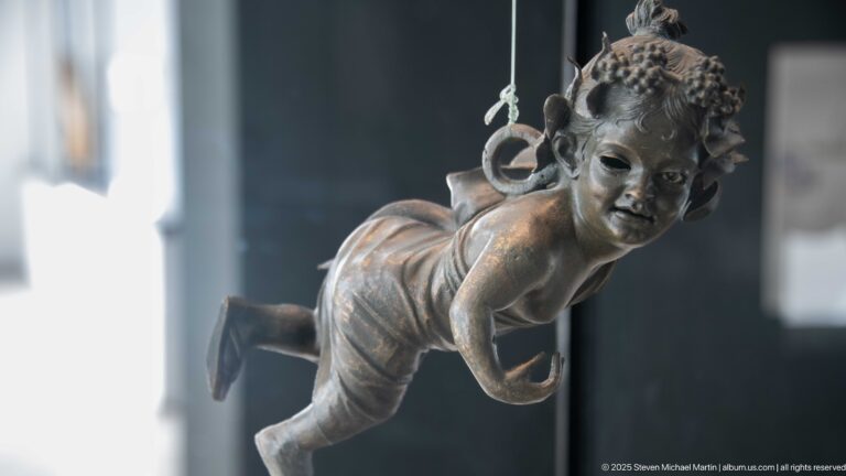Suspended bronze winged child leaning forward in flight with one leg bent and a ring behind the shoulder.