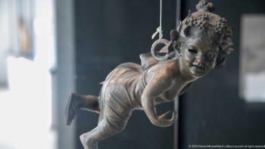 Suspended bronze winged child leaning forward in flight with one leg bent and a ring behind the shoulder.