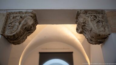 Two weathered carved stone capitals with dense leaf and volute ornament mounted beneath a horizontal stone element.