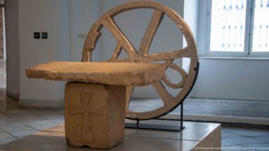 Rectangular stone slab on a carved pedestal with a cross-like motif, displayed in front of a large wooden spoked wheel.
