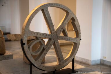 Large weathered spoked wheel with six heavy spokes, riveted rim, and central hub displayed upright on a black museum stand.