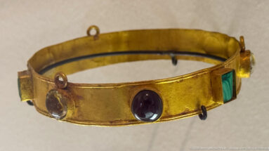 Rigid gold neck band set with oval purple and clear stones, a rectangular green stone, and several small suspension loops.