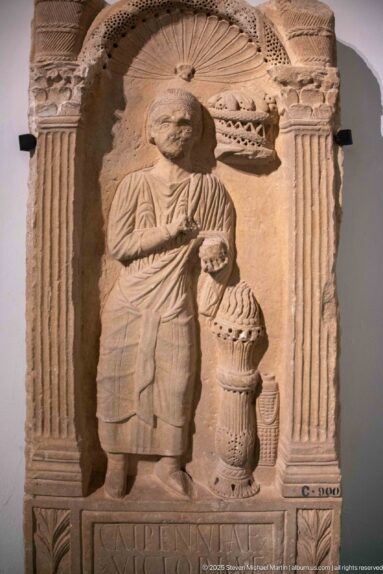 Stone funerary stele with a standing draped figure, fluted columns, curved pediment, tall urn-like vessel, and inscription below.