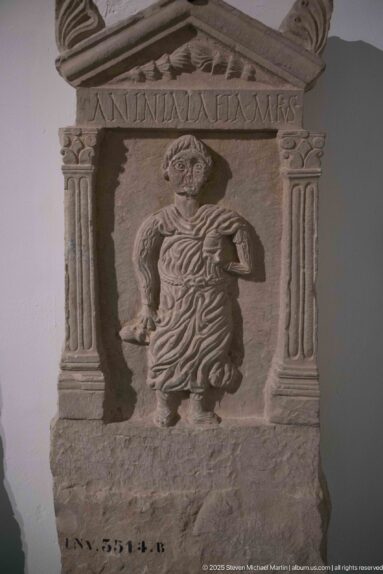 Inscribed stone stele with a standing draped woman framed by fluted columns and a triangular pediment.