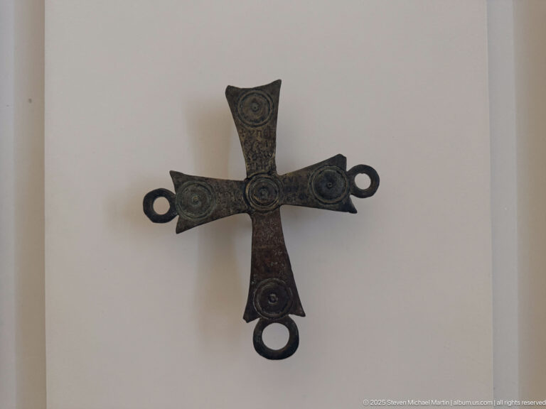 Dark bronze cross-shaped fitting with four circular medallions and ring-shaped loops projecting from three arm ends.
