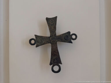 Dark bronze cross-shaped fitting with four circular medallions and ring-shaped loops projecting from three arm ends.