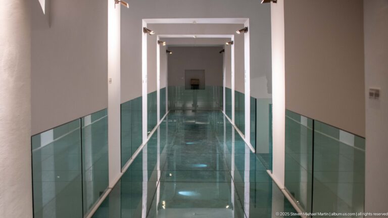 Long modern museum corridor with repeated white rectangular portals, green glass balustrades, reflective floor, and a small display at the far end.