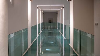 Long modern museum corridor with repeated white rectangular portals, green glass balustrades, reflective floor, and a small display at the far end.