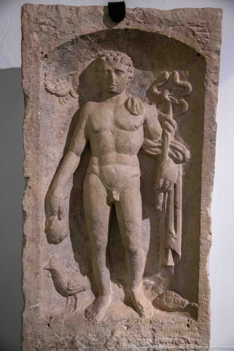 Weathered stone relief with a muscular standing man supporting a smaller figure beside a column and a small carved form at lower left.