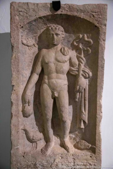 Weathered stone relief with a muscular standing man supporting a smaller figure beside a column and a small carved form at lower left.