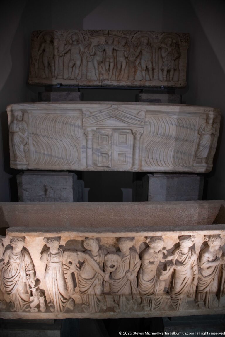 Three stacked carved marble sarcophagi with figural reliefs, strigil patterns, and architectural decoration displayed in a museum gallery.