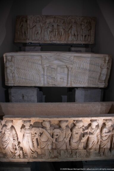 Three stacked carved marble sarcophagi with figural reliefs, strigil patterns, and architectural decoration displayed in a museum gallery.
