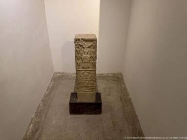 Small upright rectangular stone monument with stacked bands of geometric and symbolic carving, displayed alone on a dark base.