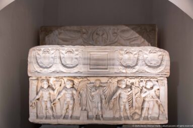 Marble sarcophagus with a row of standing carved figures below a lid decorated with looping garlands and a central rectangular panel.