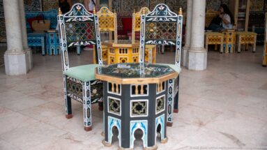 Octagonal decorated low table surrounded by tall-backed chairs painted or inlaid in turquoise, yellow, black, white, and red patterns.