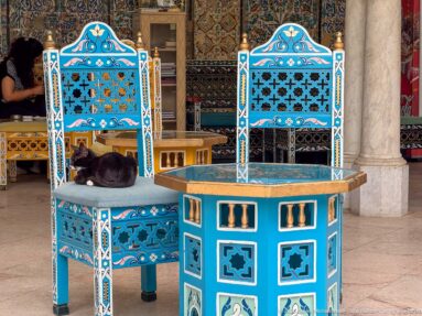 Black cat sleeping on a turquoise decorated chair beside a matching octagonal table with painted geometric panels.