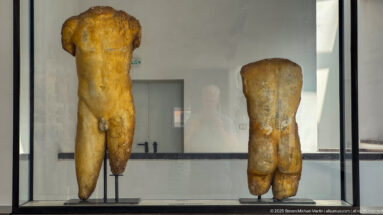 Two weathered headless stone male torsos displayed upright in a museum case.