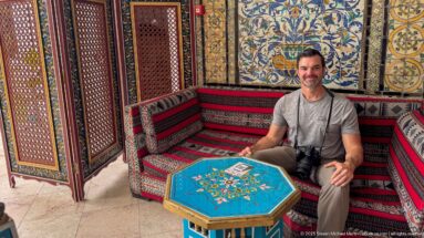 Seated male visitor with a camera in a decorated alcove with striped cushions, floral tilework, and a turquoise octagonal table.