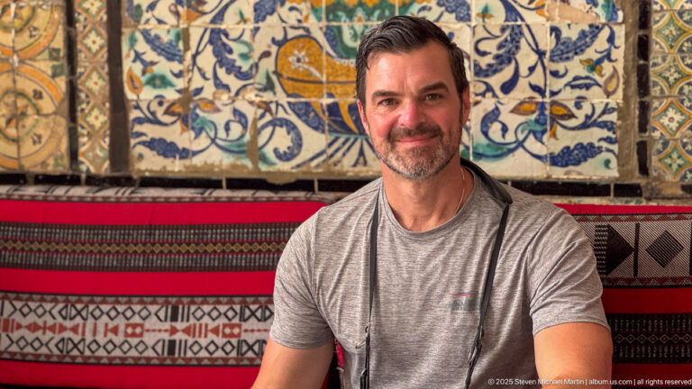 Seated male visitor in a gray shirt smiling before colorful floral tilework and red, black, and cream woven textiles.