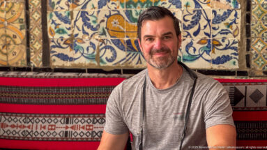 Seated male visitor in a gray shirt smiling before colorful floral tilework and red, black, and cream woven textiles.