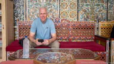 Male visitor seated on a red bench holding a phone, surrounded by painted wood furniture, patterned tilework, and a decorated table.