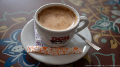 White cup of coffee with crema on a saucer, spoon, and sugar packet resting on a colorful decorated tabletop.